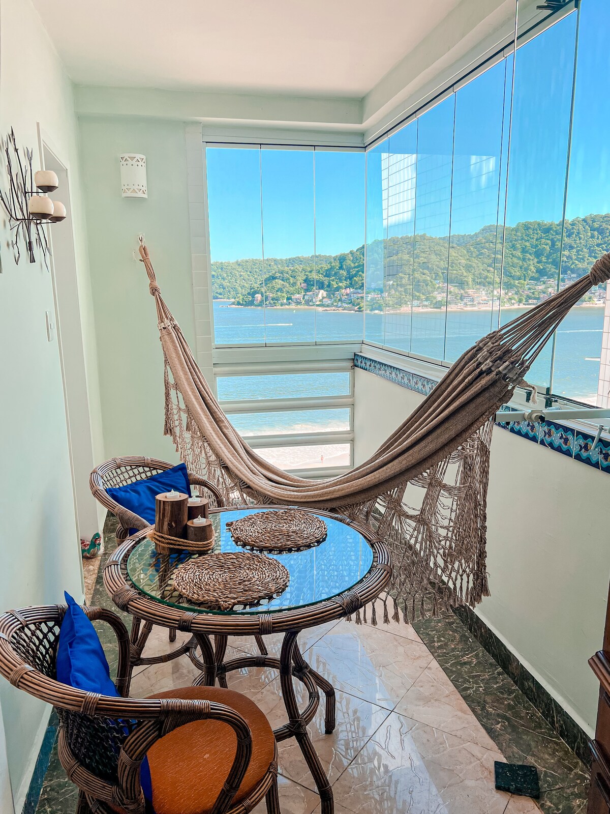 A cozy balcony space features a hammock suspended between two walls, inviting relaxation. A small round table is set with woven placemats and has two chairs. Panoramic glass windows frame a stunning view of the beach and coastline, allowing natural light to fill the area.