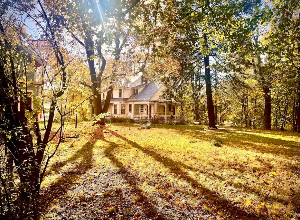 The Victorian mansion is surrounded by vibrant fall foliage, with sunlight filtering through the trees. The lawn is covered in a blanket of fallen leaves, and the long shadows cast by the trees create a serene atmosphere. The house features a welcoming porch and gently sloped roof.