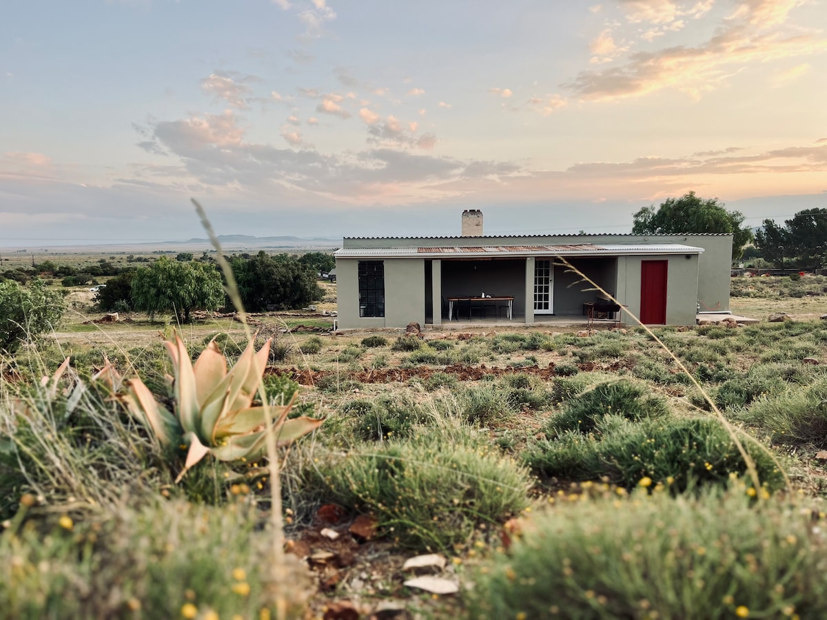 The cottage is situated amidst a vast landscape, featuring a combination of low vegetation and a few flowering plants. The structure has a red door and simple design, with a veranda visible at the front. The sky displays soft colors at sunset, enhancing the serene setting.