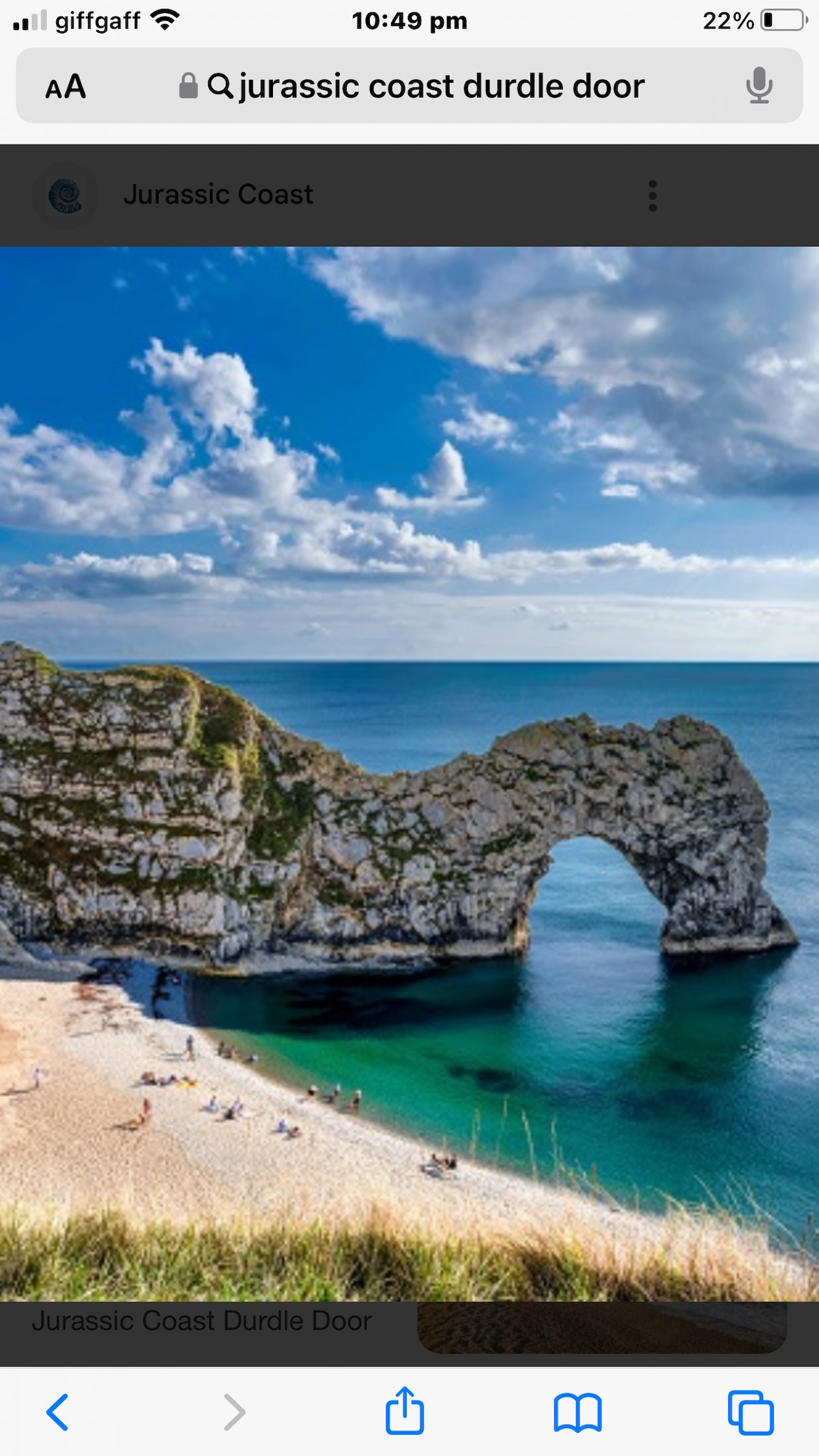 The image features the iconic Durdle Door, a natural limestone arch, set against a backdrop of a clear blue sky with scattered clouds. The turquoise sea gently meets the sandy beach, where a few people can be seen enjoying the shore.