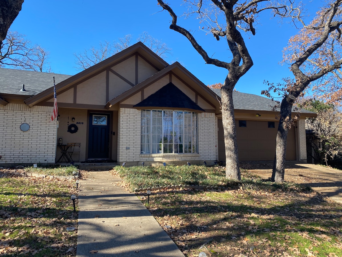 The front exterior of the studio is showcased, featuring a light-colored brick façade and a sloped roof. A welcoming entrance is highlighted by a dark door and large windows with grid patterns, surrounded by mature trees and a manicured walkway.