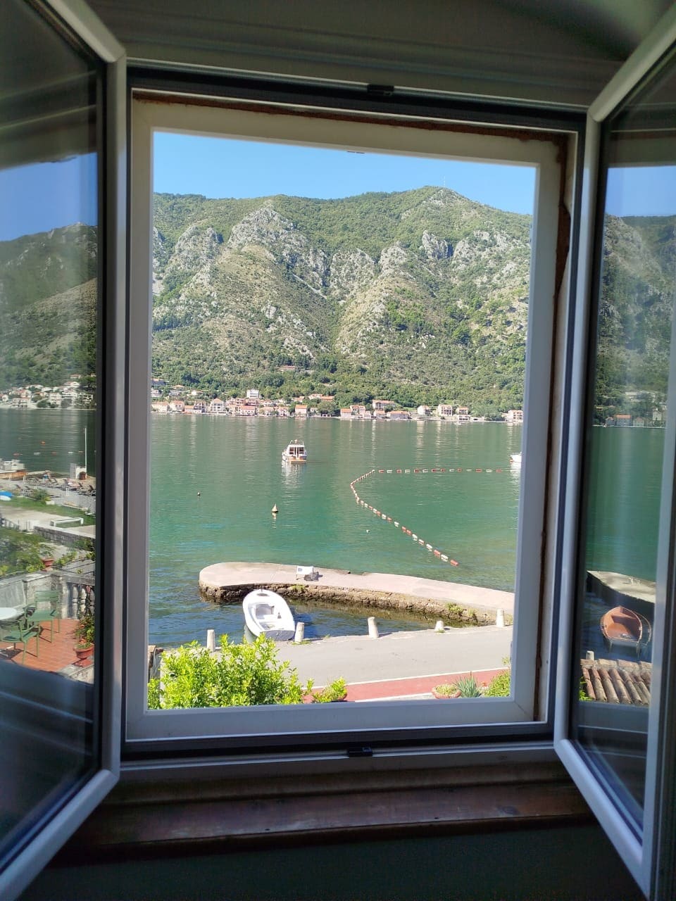 A serene view is captured through an open window, revealing calm waters of the bay reflecting the surrounding mountains. Various boats can be seen gently bobbing in the harbor, while quaint buildings line the shoreline, framed by lush greenery in the backdrop.