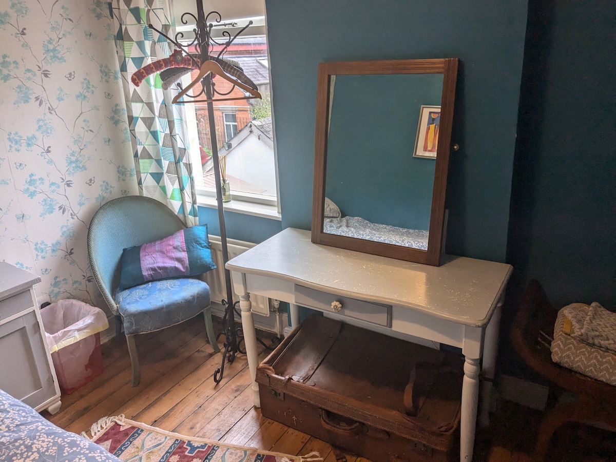 A simple room features a wooden dressing table with a mirror above it, accompanied by a vintage suitcase and a blue upholstered chair. A decorative coat rack stands next to the window, while patterned wallpaper adds depth to the calming atmosphere.