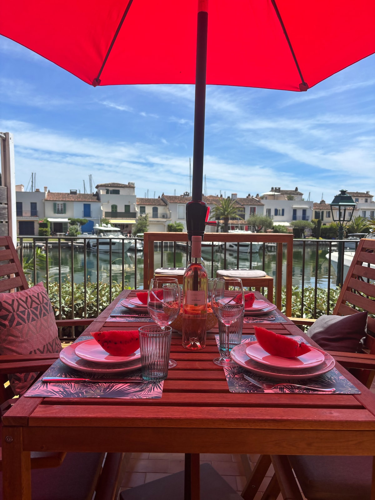 A dining table set for four is displayed under a vibrant red umbrella, overlooking a canal. The table is adorned with plates, glasses, and a bottle of rosé, offering a view of charming buildings and boats in the background.