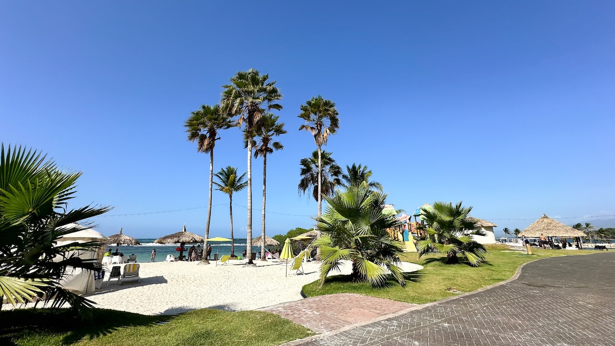 A sandy beach area is depicted, featuring several palm trees swaying in the gentle breeze. Beach umbrellas provide shade on the white sand, while sun loungers are arranged nearby. A clear blue sky complements the tranquil atmosphere by the water's edge.