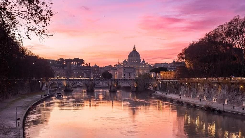 An evening view of the Tiber River reflects the soft pink and purple hues of the sunset. The silhouette of St. Peter's Basilica is prominently featured in the background, framed by the trees lining the riverbank. The peaceful waters invite a sense of tranquility.