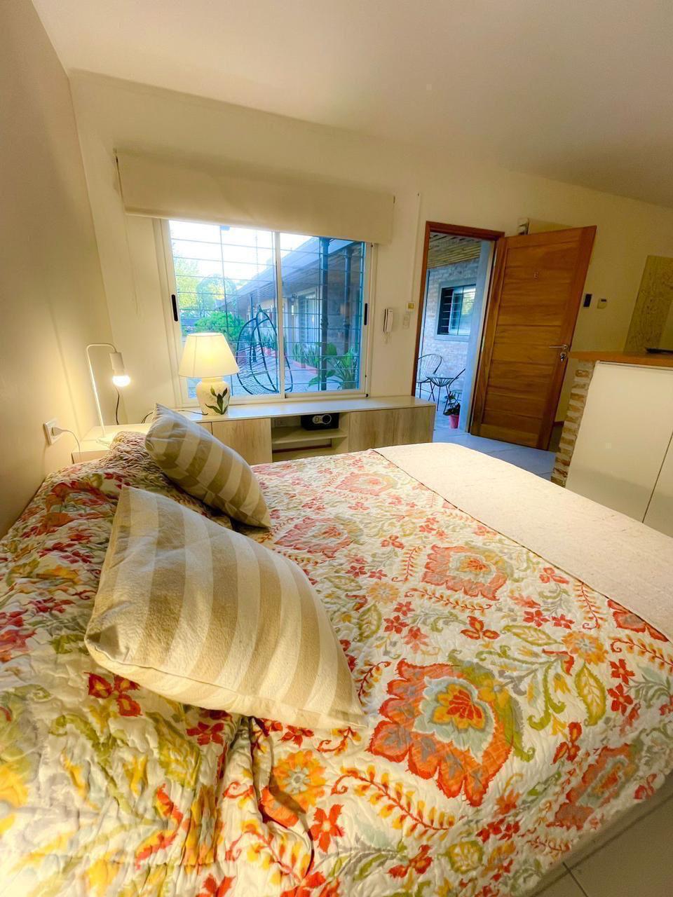 A well-lit bedroom features a spacious bed adorned with a colorful quilt and decorative pillows. Natural light enters through a large window, with a view of greenery. A bedside lamp and a wooden surface add to the room's functionality, enhancing the cozy ambiance.
