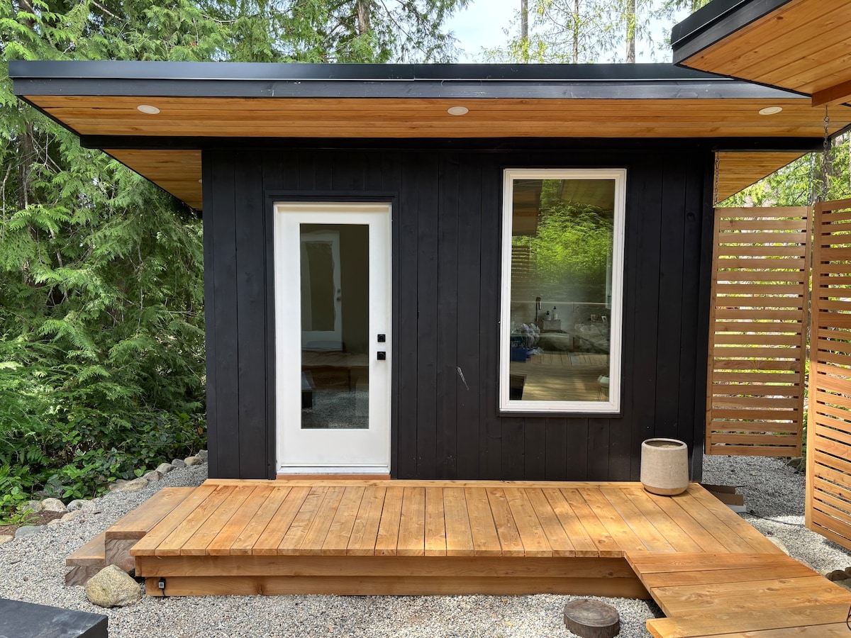The exterior of a modern wooden unit is shown, featuring a blend of dark wood and natural elements. A wooden deck surrounds the unit with a smooth transition to gravel. Large windows provide glimpses of natural light and surrounding greenery.