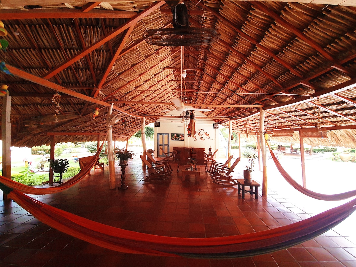 A spacious common area features a high, thatched roof supported by wooden beams. Several rocking chairs are arranged around a central wooden table. Colorful hammocks hang between posts, inviting relaxation, and plants are strategically placed throughout the space, enhancing the tranquil atmosphere.
