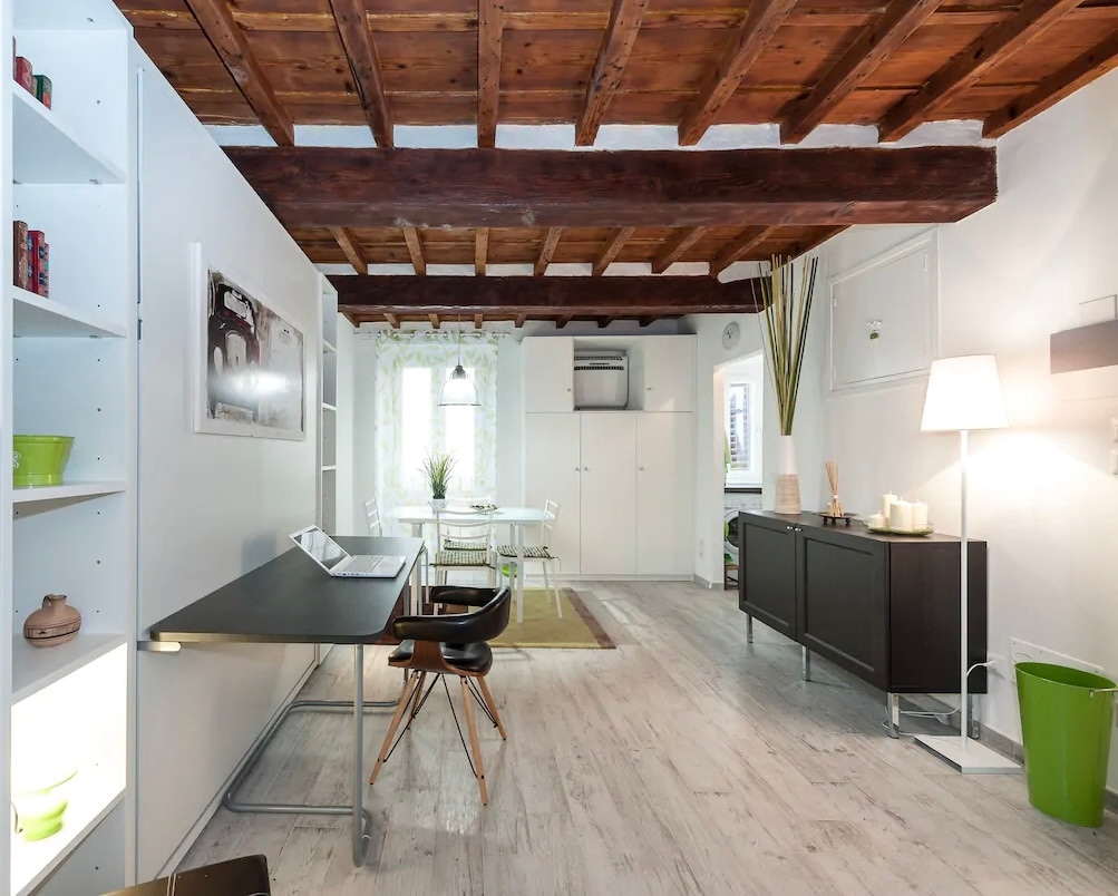 An open living space with high wooden beam ceilings is shown. A minimalist desk is positioned on the left, accompanied by a chair. To the right, a dining area is highlighted by a white table and four chairs. Natural light enters through a window, enhancing the bright decor.