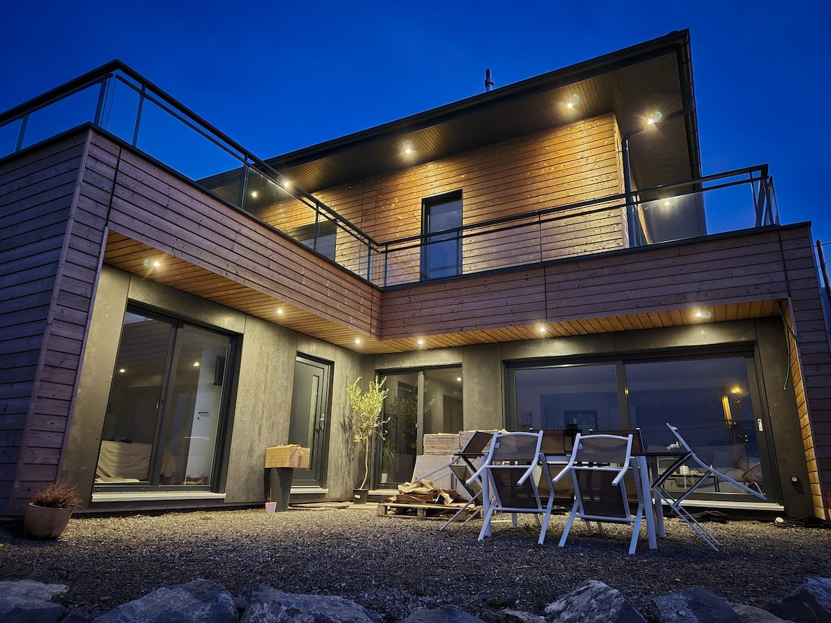 The modern exterior features a wooden façade and large glass doors, revealing a sophisticated structure. Outdoor seating is provided on a gravel terrace, with several lounge chairs positioned for relaxation. Soft lighting enhances the evening ambiance, inviting guests to unwind.