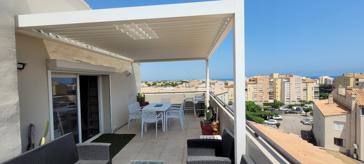 A spacious terrace features a covered dining area with a table and chairs, surrounded by potted plants. The view extends towards the horizon, showcasing nearby buildings and a glimpse of the sea. Comfortable seating arrangements provide a relaxing space for gatherings.