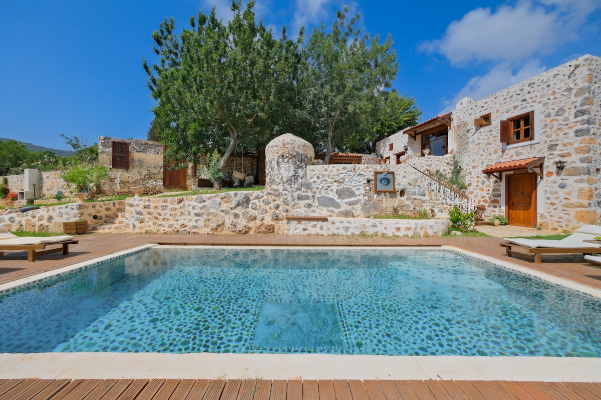 A natural stone swimming pool is surrounded by wooden decking and lounge chairs, offering a serene space for relaxation. Lush greenery and trees frame the pool area, while historical stone structures provide a rustic backdrop, contributing to the tranquil setting.