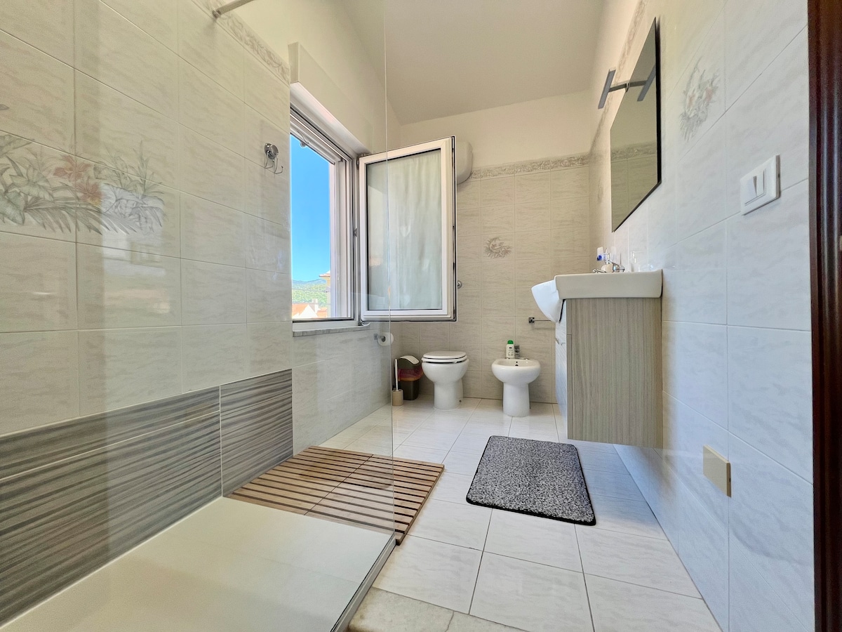 A well-lit bathroom features a walk-in shower with a tiled floor and a wooden mat. A window allows natural light, and a mirror is positioned above the sink. Storage space is provided by a wooden cabinet, with a toilet and a rug occupying the floor area.