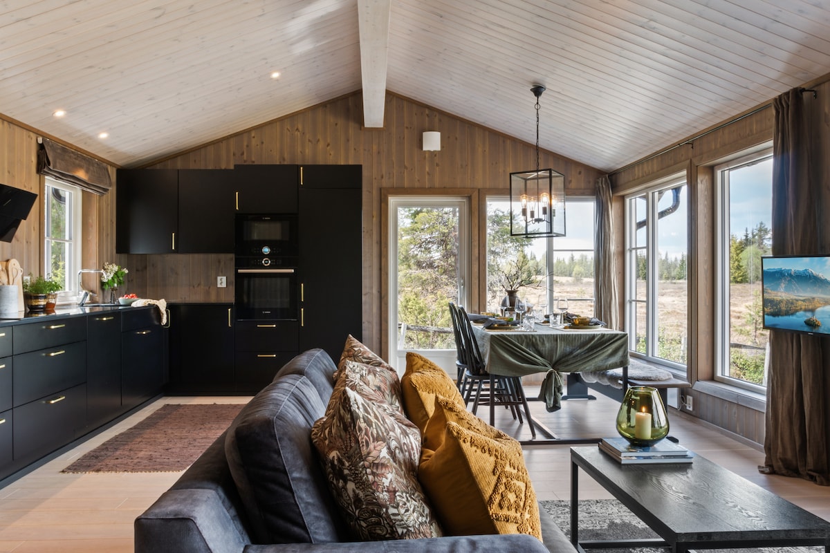 An open-plan living area featuring a modern kitchen with sleek black cabinetry. Large windows provide natural light and views of the surrounding landscape. A comfortable seating arrangement is positioned nearby, accented with textured cushions. A dining table is set at the opposite end, enhancing the inviting space.