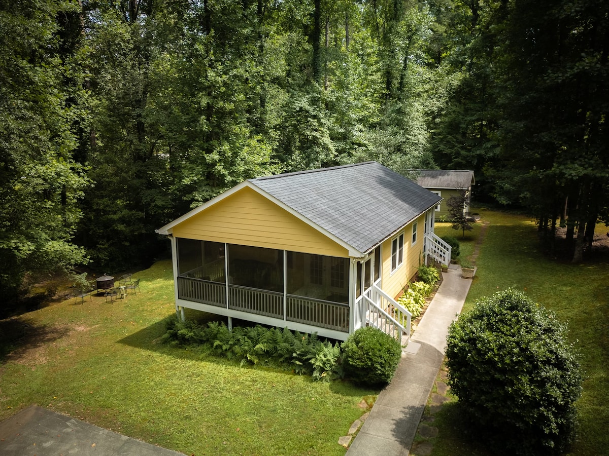 The cabin is surrounded by lush trees, with a bright yellow exterior and a spacious screened-in porch. A pathway leads to the entrance, while a table and chairs are visible in the grassy area. The serene environment creates a peaceful setting.