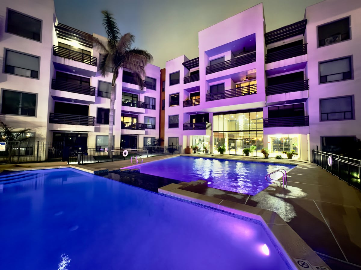 A modern building is illuminated at night, showcasing a large resort-style pool with calming blue water. Lush tropical plants are strategically placed around the pool area, and glass doors lead into the inviting lobby, enhancing the overall aesthetic of the outdoor space.