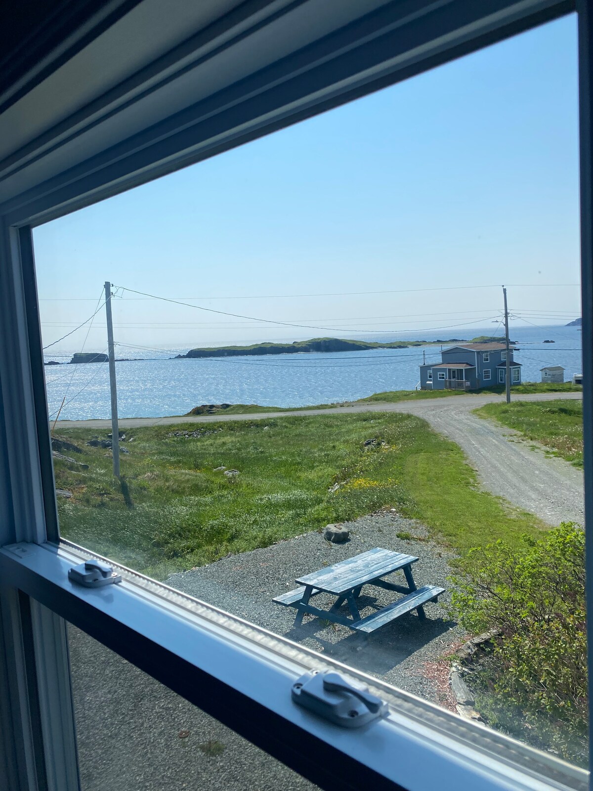 A view from the window captures a serene landscape featuring rolling green grass and a gravel pathway leading toward the Atlantic Ocean. The distant Ragged Islands and a quaint blue house are visible, framed by a clear blue sky.