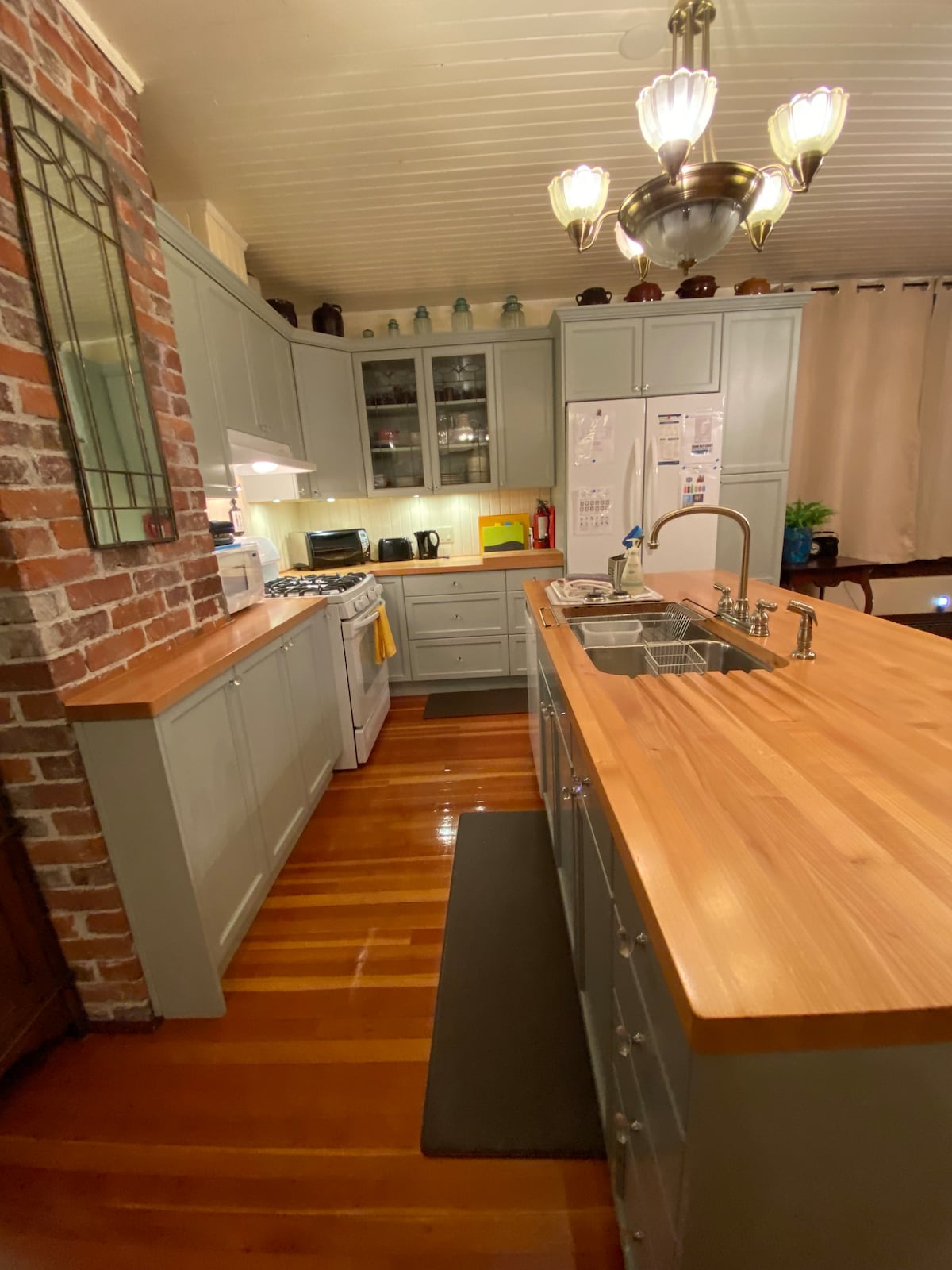 A fully equipped kitchen features soft green cabinetry with a wood countertop. The space is illuminated by overhead lights and a decorative fixture. Appliances include a stove and refrigerator, with dishes and utensils visible on the shelves. A rug is placed in front of the sink area.