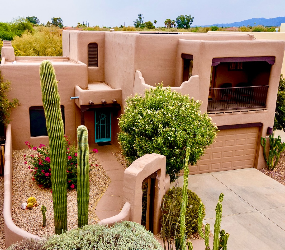 The house features a classic adobe style exterior, surrounded by desert landscaping. Tall saguaro cacti and colorful bougainvillea are seen in the front yard, while the balcony offers views of the surrounding landscape. A driveway leads to the garage, showcasing the home's unique architecture.