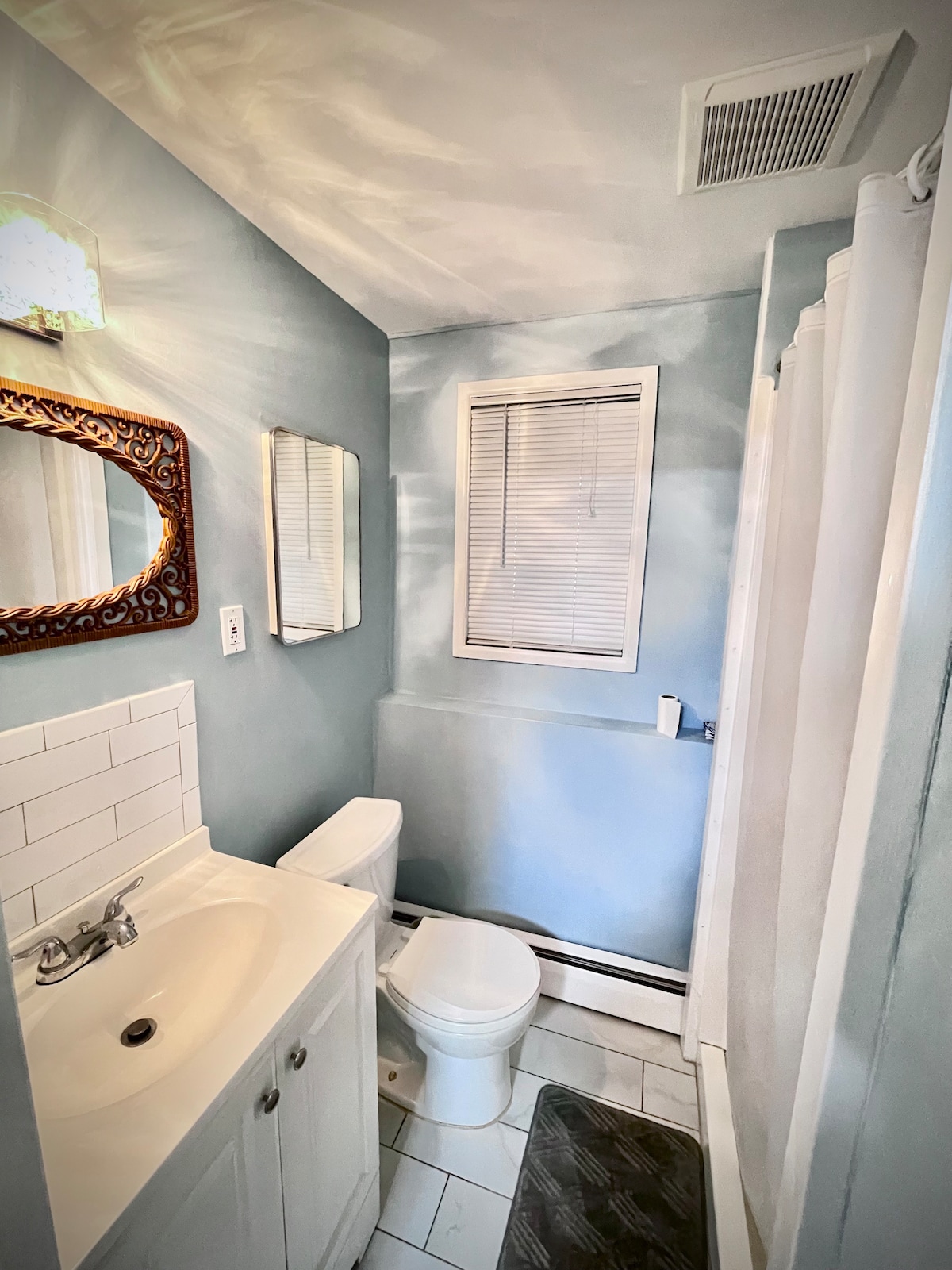 A well-lit bathroom features soft blue walls and white tiles. A small sink with a classic faucet is positioned next to a modern toilet. Natural light enters through a window covered with horizontal blinds. A framed mirror hangs above the sink, adding a decorative touch.