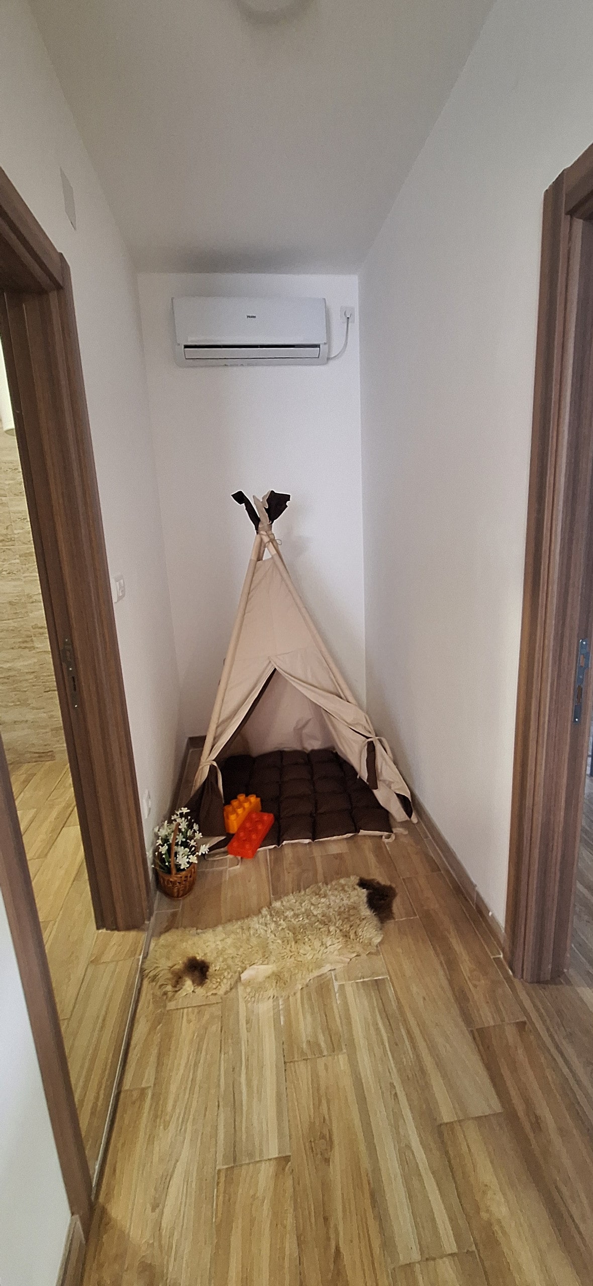 A cozy corner is highlighted by a small, fabric teepee with a soft, cushioned interior. Nearby, a textured rug adds warmth to the space. An air conditioning unit is mounted on the wall, and light-colored wooden flooring creates a serene atmosphere.