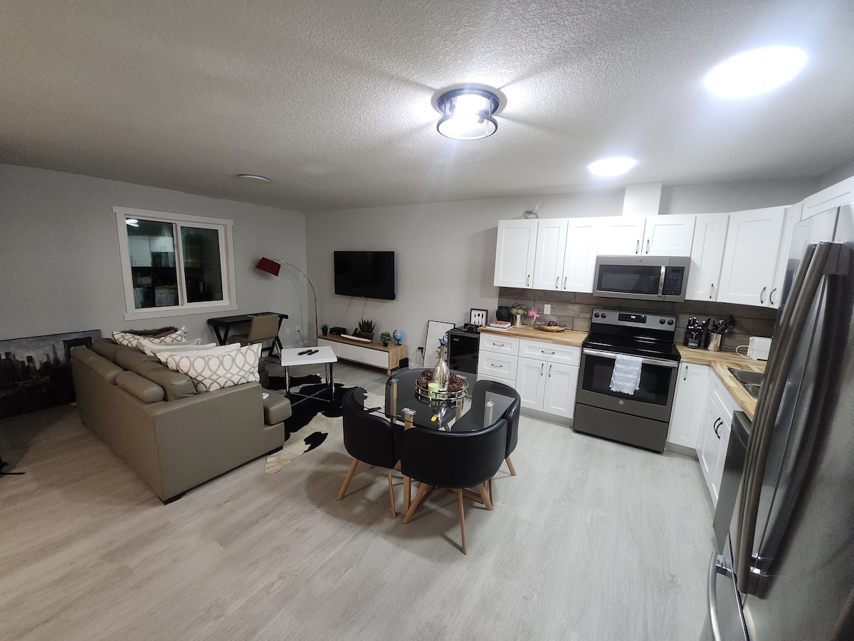 An open-plan living space showcases a modern kitchen and cozy seating area. A sleek stainless steel appliance set complements white cabinetry. A dark dining table, surrounded by four chairs, is positioned near a comfortable sofa, creating an inviting area for relaxation and dining.