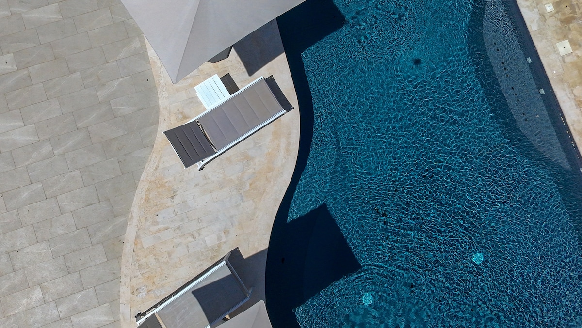 A clear view of a swimming pool, featuring gently curving edges and surrounded by a spacious stone deck. Two sun loungers with reclining backs are positioned nearby, offering a comfortable space to relax. A shaded area is present, providing relief from the sun.