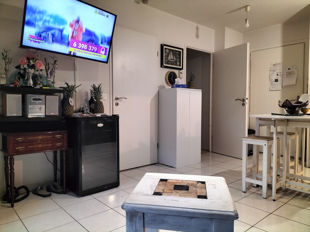 A living area is presented with tiled flooring and a mix of furnishings. A wall-mounted television displays a program. A small table with a decorative centerpiece sits in front of a compact refrigerator, while wooden stools surround a dining table against a plain wall.