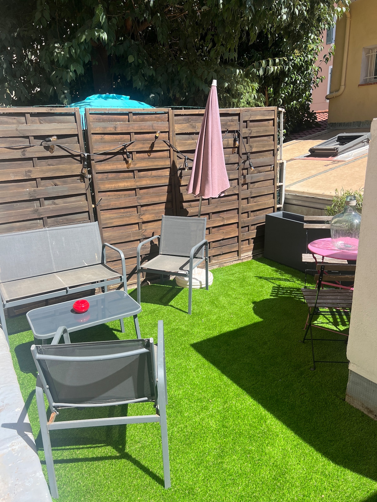 A private terrace is equipped with two chairs and a glass-topped table, surrounded by a wooden fence. Artificial grass covers the floor, while a pink umbrella provides shade. A small grill is also visible, creating an inviting space for outdoor relaxation.