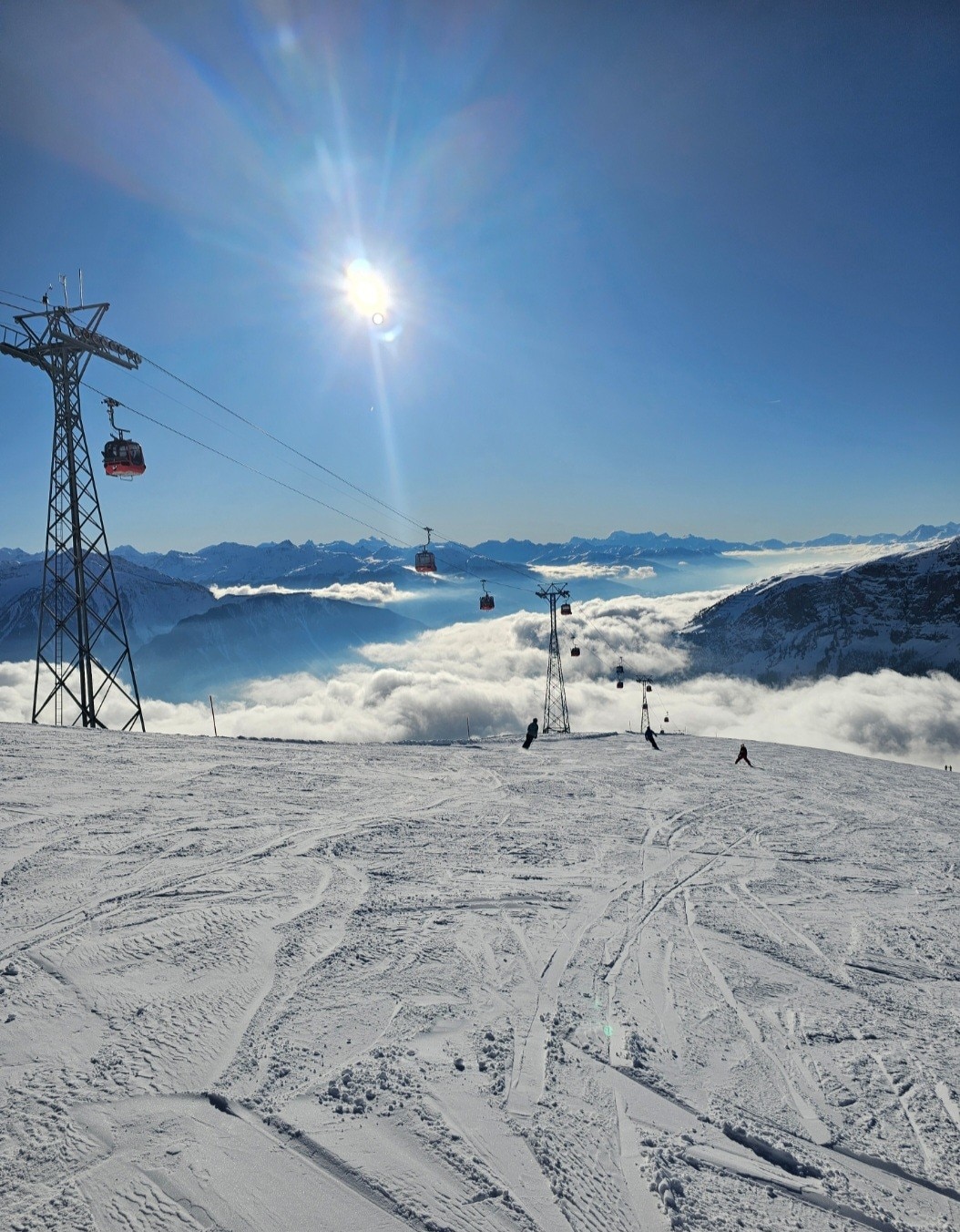 A panoramic view of snow-covered slopes is seen under bright sunlight, with ski lift gondolas ascending against a backdrop of mountains and clouds. The clear blue sky complements the winter landscape, inviting outdoor enthusiasts to explore the area.