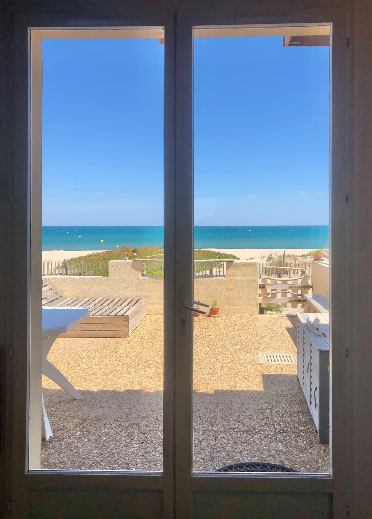 A view through glass doors reveals a serene beach with soft sand and calm blue waters. The outdoor space features a wooden seating area and potted plants, framed by a clear sky. The scene embodies a relaxed coastal atmosphere, inviting moments of tranquility.