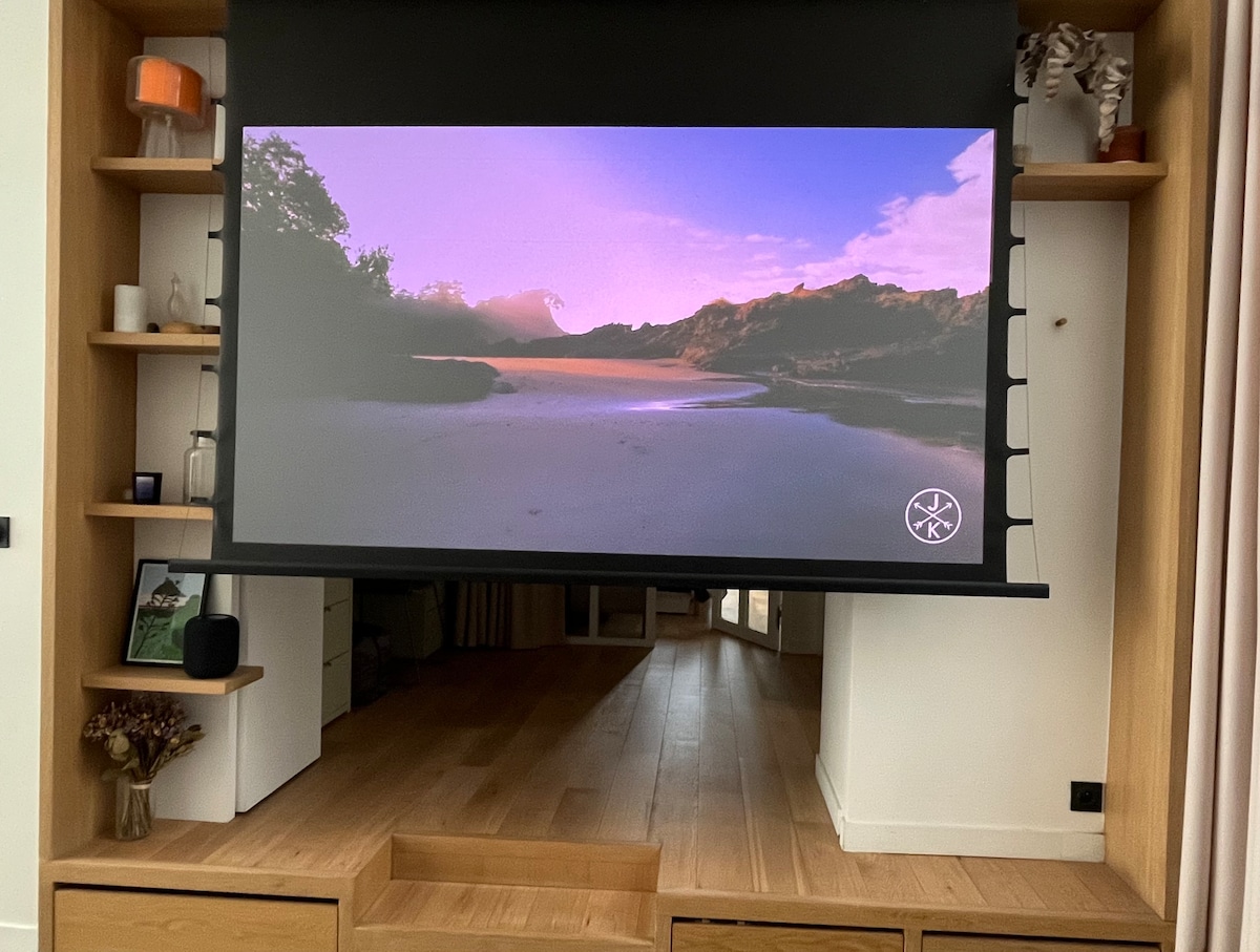A large projection screen is lowered in a living area, displaying a tranquil landscape scene with mountains and a calm body of water under a purple sky. The surrounding wooden shelves contain decorative objects, contributing to a modern aesthetic.
