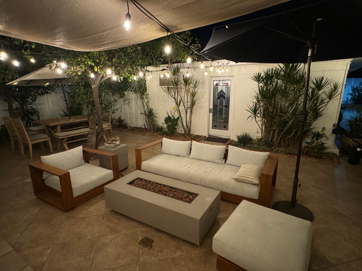 A cozy outdoor patio area is illuminated by soft string lights, featuring a modern seating arrangement with two couches and a coffee table centered around a fire pit. An umbrella provides shade, while tropical plants enhance the tranquil setting.
