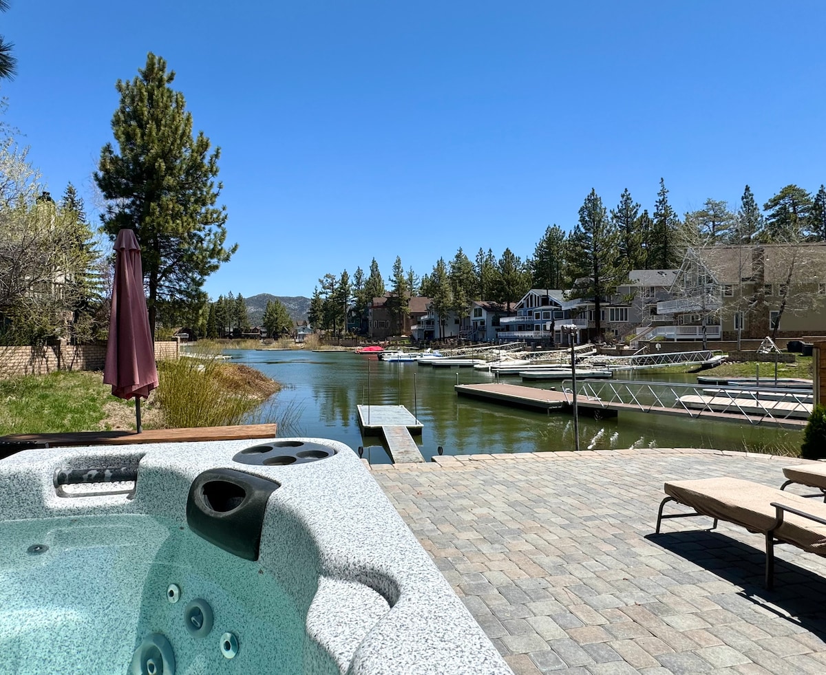 A private hot tub is positioned on a stone patio, overlooking a serene lake. The view captures the calm water surrounded by trees and homes, with docks visible along the shoreline. Sunlight brightens the scene, enhancing the inviting outdoor space.