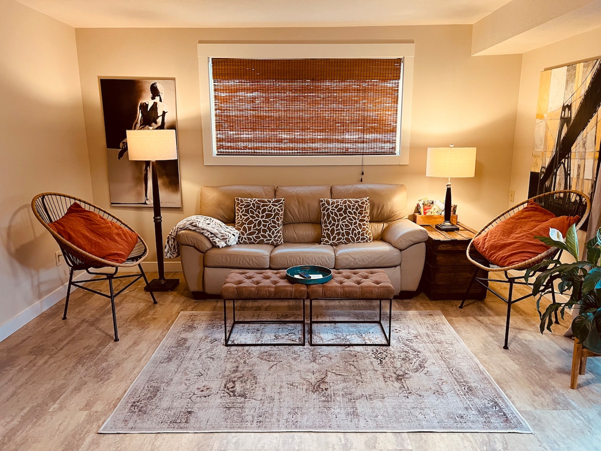 The living area features a soft brown sofa adorned with patterned cushions, situated on a neutral-toned area rug. Two stylish chairs with orange accents flank the sofa. Warm light is provided by two lamps, enhancing the cozy atmosphere of the room.