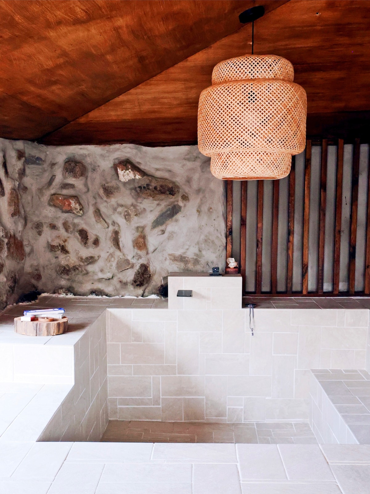 A serene indoor bathing area features natural stone walls and textured wooden ceiling. The space includes a sunken soaking tub and a wooden stool, complemented by a large woven pendant light that adds warmth to the environment.
