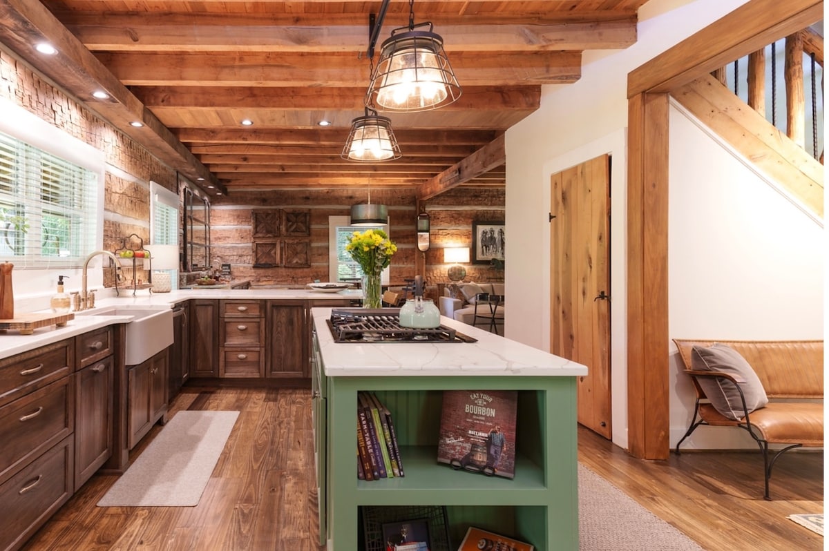 An inviting kitchen showcases a large central island with a green base, complemented by wooden cabinetry and modern appliances. Exposed wooden beams create a cozy atmosphere, while natural light flows through the windows, highlighting decorative elements on the counter and shelves.