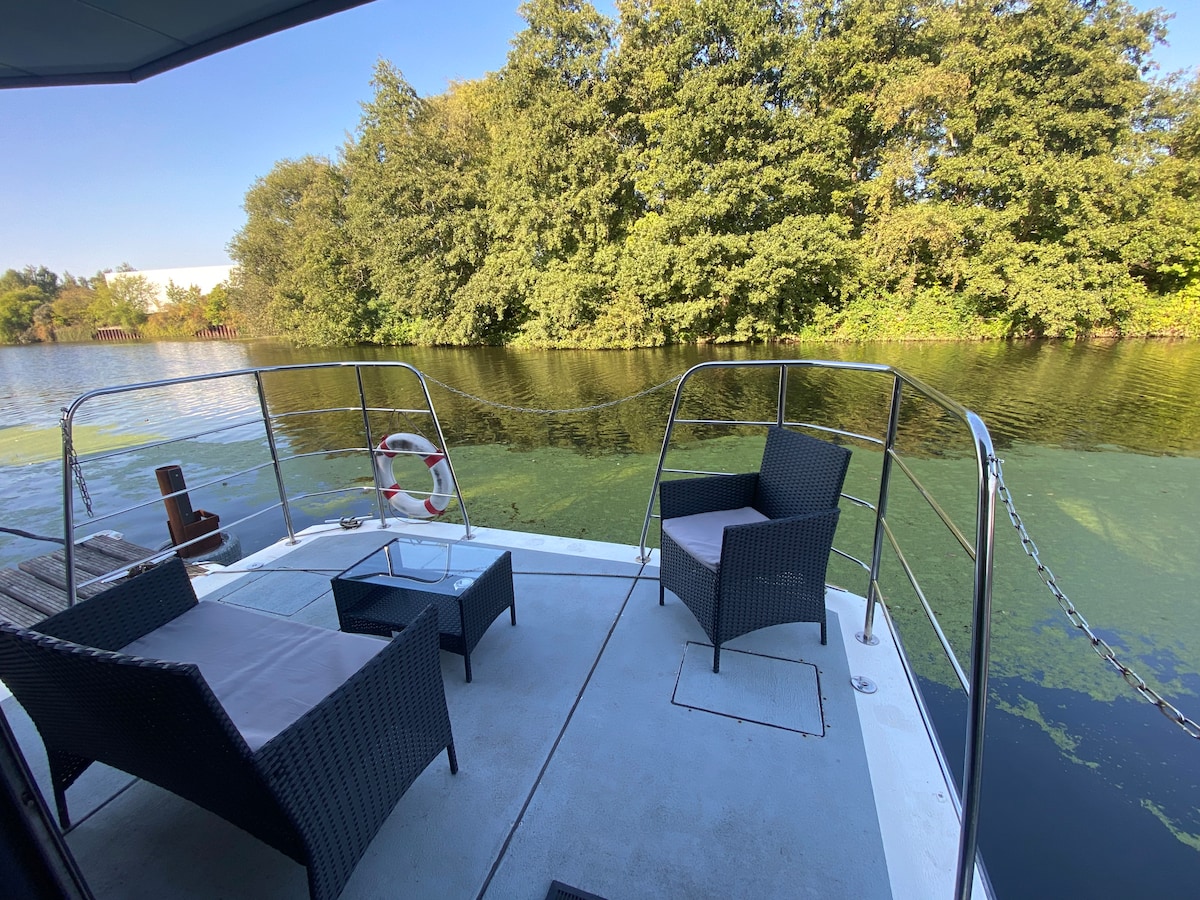 An outdoor terrace is depicted, featuring two wicker armchairs and a small table. Surrounding greenery reflects in the calm water, creating a serene environment. A safety buoy is visible on the side, and a clear blue sky enhances the setting.