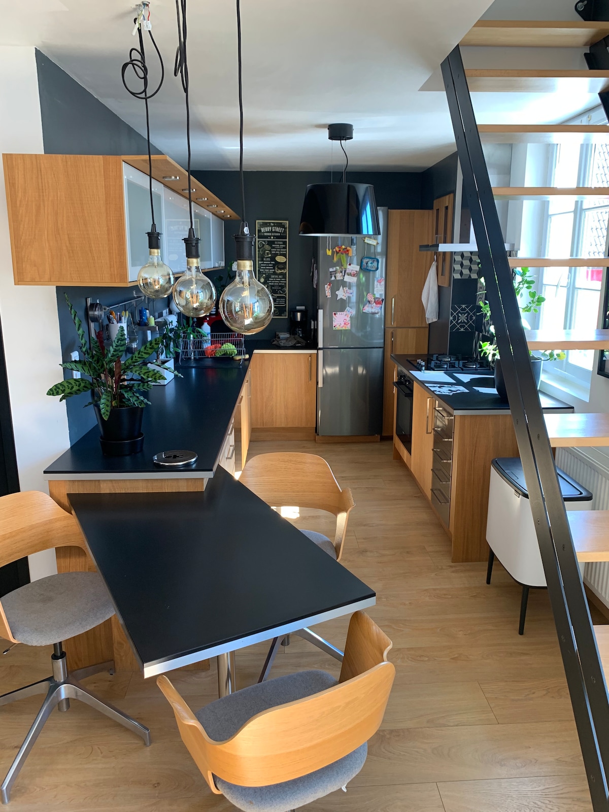 A modern kitchen space is highlighted by wood cabinetry and sleek black countertops. A dining table with four light wood chairs is positioned nearby. Warm light is provided by hanging glass pendant lamps. The atmosphere is enhanced by greenery placed on the countertop.