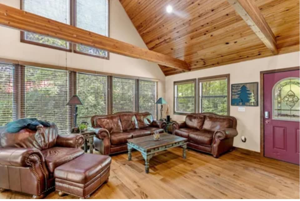 A comfortable living area features leather sofas and a plush armchair, arranged around a wooden coffee table. Large windows provide natural light and views of the surrounding greenery, while wooden beams add warmth to the high ceiling.
