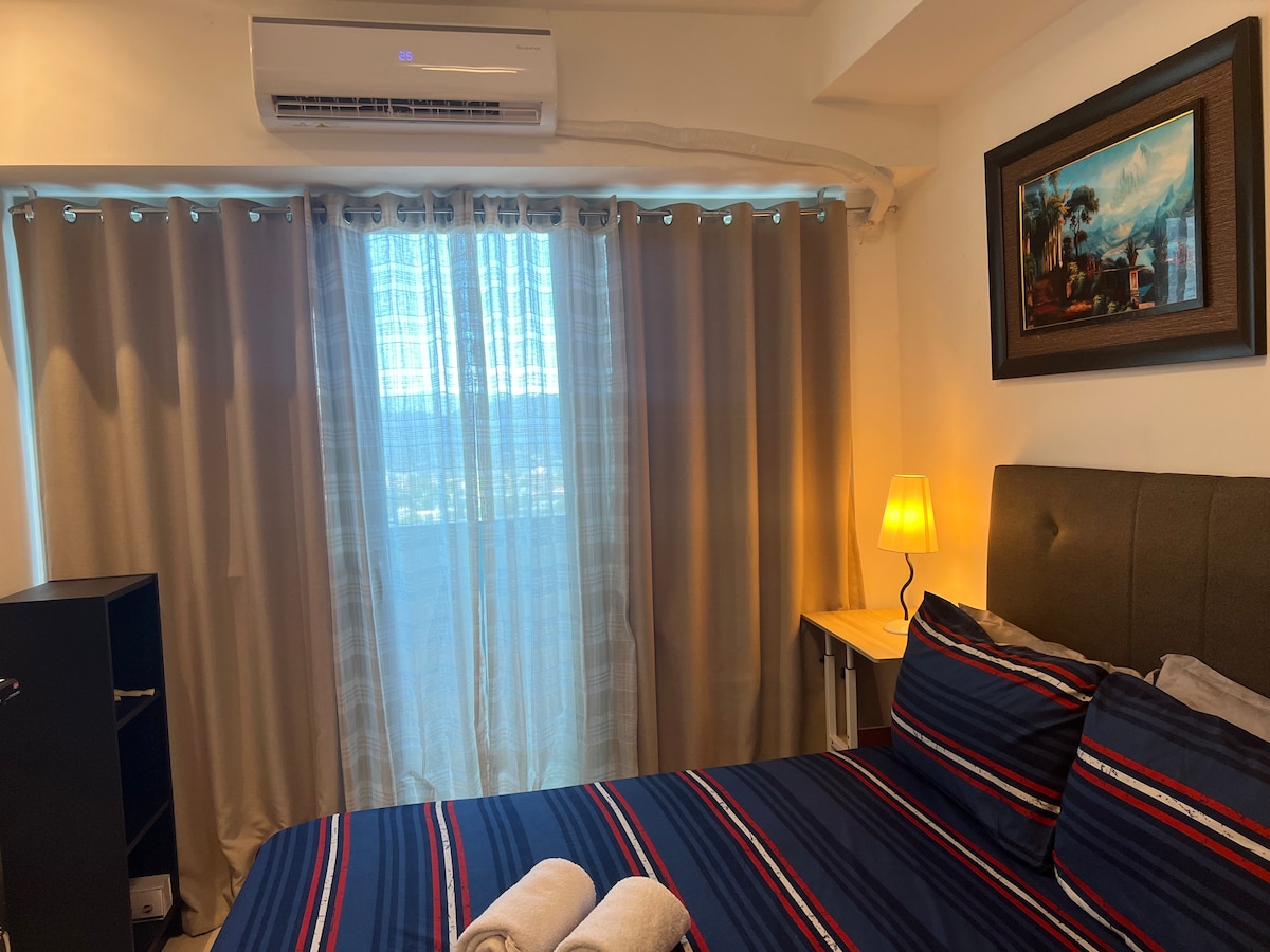 A cozy sleeping area features a spacious bed adorned with striped bedding and neatly arranged towels. Soft curtains filter natural light from a nearby window, while a lamp provides warm illumination. A painting with a landscape adds a touch of artistry to the wall.