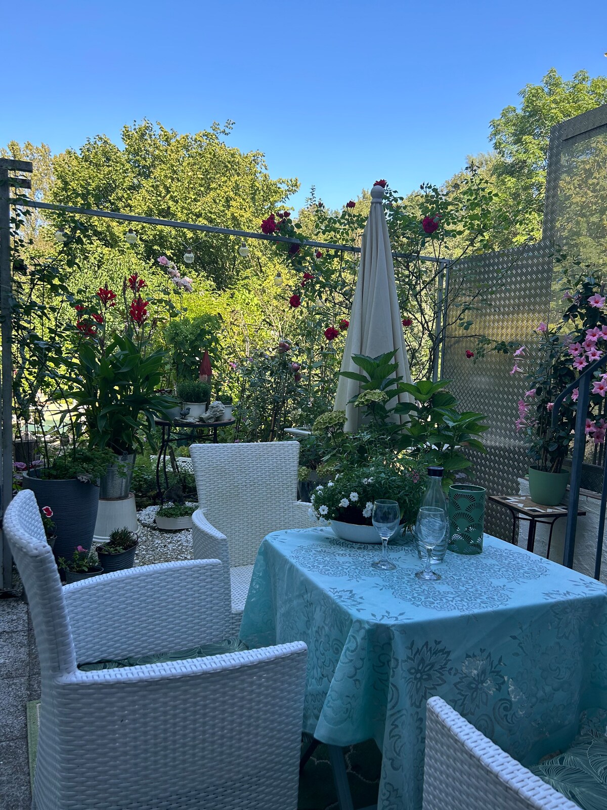 A small outdoor balcony is adorned with lush greenery and blooming flowers in various pots. A table covered with a light blue cloth is set for two, accompanied by white wicker chairs. An umbrella provides shade, enhancing the cozy atmosphere.