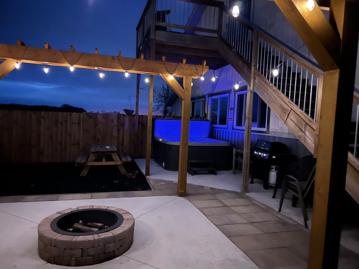An inviting outdoor area is illuminated by string lights, featuring a fire pit surrounded by paved stones. A hot tub sits nearby, complemented by a picnic table and grill, with a wooden pergola supporting the lights, creating a cozy atmosphere for evening relaxation.