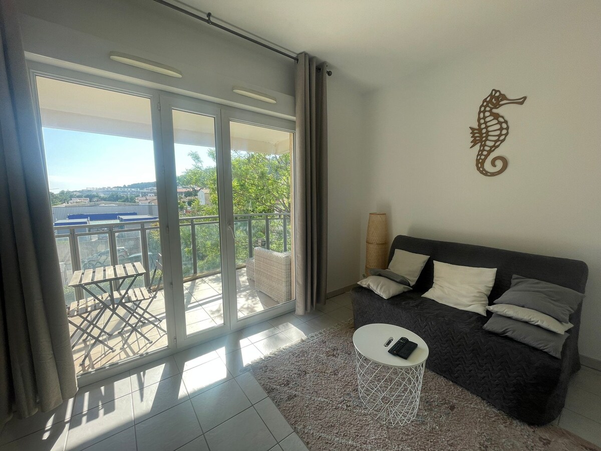 A bright living area is shown with large glass doors leading to a balcony, allowing natural light to fill the space. A gray sofa is displayed with decorative cushions. A small round table and two chairs are visible on the balcony, surrounded by greenery.