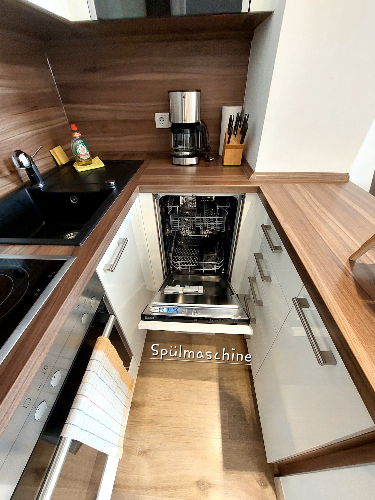 A modern kitchen space features sleek wooden countertops and a black sink. An open dishwasher is displayed, with minimalist cabinetry above. A coffee maker and dish soap are positioned nearby, highlighting the functional layout of the kitchen area.