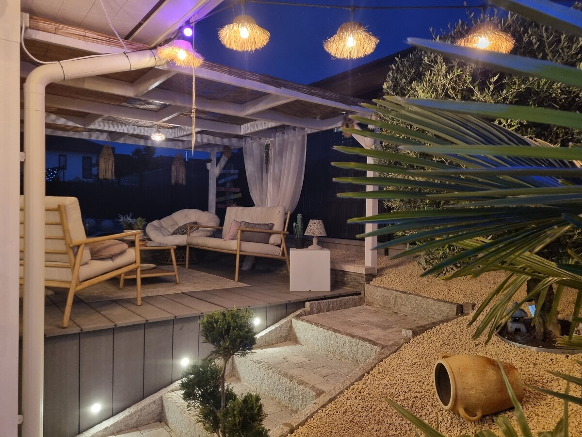 An outdoor lounge area is set beneath a covered terrace, featuring comfortable seating and ambient lighting. Steps lead down to a landscaped garden with pebbles and lush greenery. Decorative elements like a ceramic planter are visible, enhancing the serene environment.