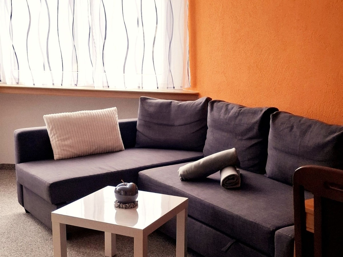 A modern seating area features a dark blue sectional couch positioned near a window with light curtains. A small white coffee table rests in front, accented by decorative items. A folded blanket and a cushion are casually placed on the couch.