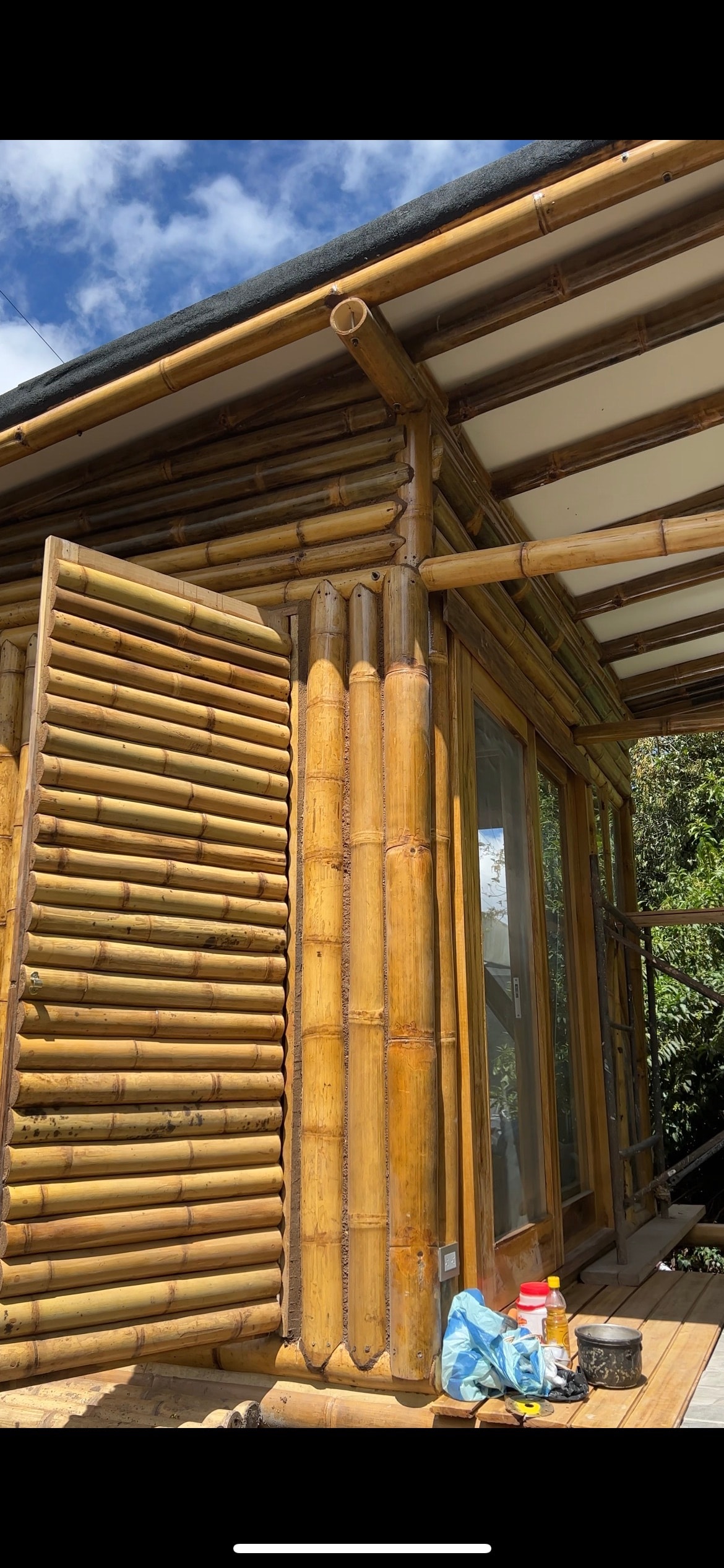 A structure constructed with bamboo showcases a combination of natural textures and modern design. Large glass windows allow light to flood the interior, while detailed bamboo siding enhances the aesthetic. The roof features wooden beams, contributing to a rustic appearance.