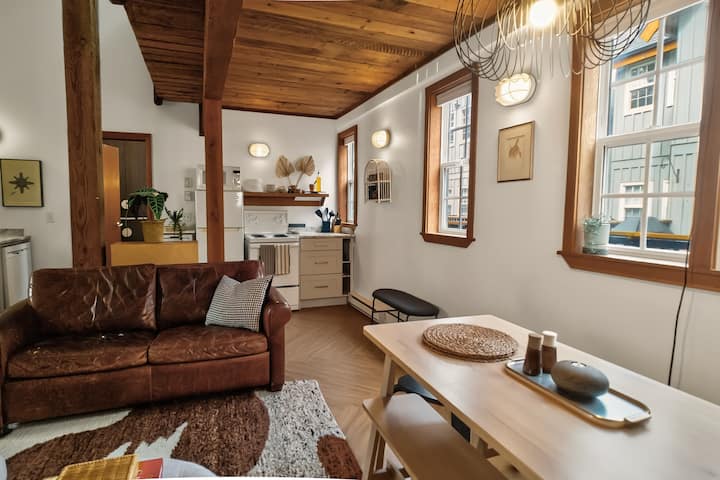 Sala con un sofá de cuero marrón, un juego de comedor minimalista y una zona de cocina a la vista